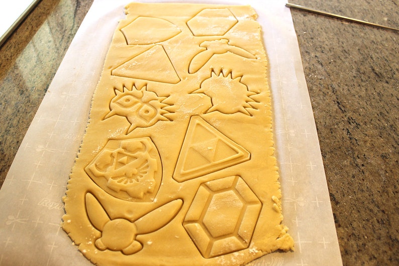 Puede incluir: Una hoja de masa para galletas con varias formas cortadas, incluyendo un escudo, una espada, un tri&aacute;ngulo, un diamante y una estrella. La masa es de color marr&oacute;n claro y est&aacute; sobre papel vegetal blanco.