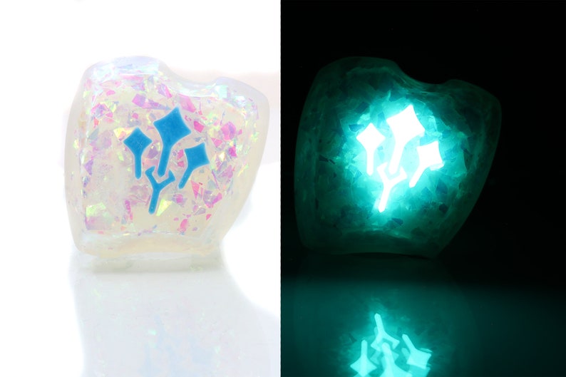 K&ouml;nnte beinhalten: Ein wei&szlig;er, irisierender W&uuml;rfel aus Harz mit einem blauen, dreispitzigen Sternensymbol. Der W&uuml;rfel leuchtet im Dunkeln gr&uuml;n.
