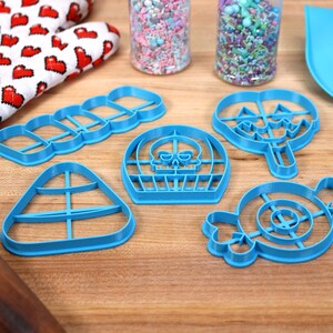 May include: A set of six blue plastic cookie cutters shaped like Halloween candy. The cutters are on a wooden surface with a rolling pin, a spatula, and two jars of sprinkles.