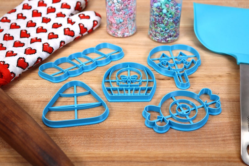 May include: A set of six blue plastic cookie cutters shaped like Halloween candy. The cutters are on a wooden surface with a rolling pin, a spatula, and two jars of sprinkles.