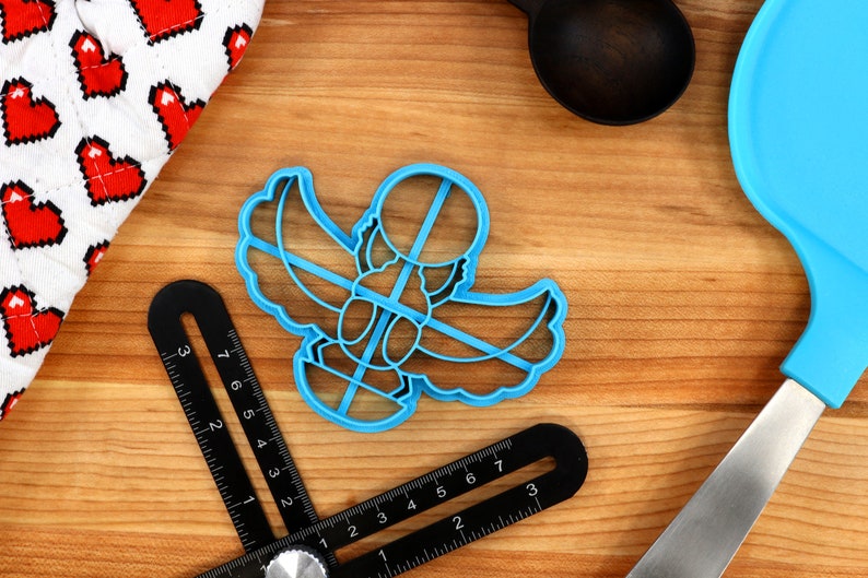 May include: Blue plastic cookie cutter shaped like a scarab beetle with wings. The cutter is on a wooden surface with a ruler and a blue spatula.