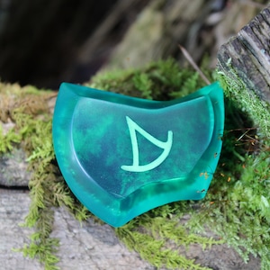 May include: A teal green, semi-circular, resin token with a white symbol of a stylized arrow. The token is sitting on a mossy log.