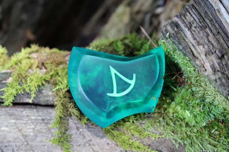 May include: A teal green, semi-circular, resin token with a white symbol of a stylized arrow. The token is sitting on a mossy log.