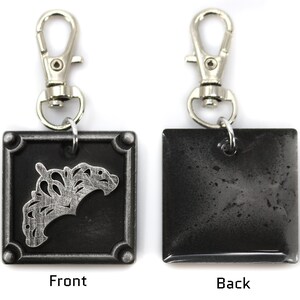 May include: A silver and black metal keychain with a square charm. The front of the charm features a silver crown design. The back of the charm is plain.