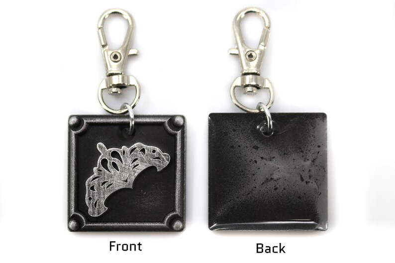 May include: A silver and black metal keychain with a square charm. The front of the charm features a silver crown design. The back of the charm is plain.