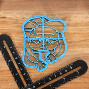 May include: A blue plastic cookie cutter shaped like a cartoon character with long hair, wearing a hat and a sad expression.