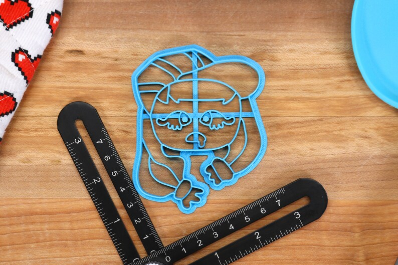 May include: A blue plastic cookie cutter shaped like a cartoon character with long hair, wearing a hat and a sad expression.