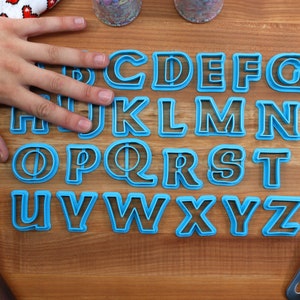 May include: A set of 26 blue plastic alphabet cookie cutters on a wooden surface. The cutters are arranged in alphabetical order.