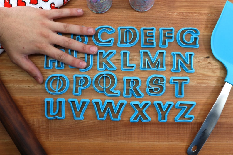 May include: A set of 26 blue plastic alphabet cookie cutters on a wooden surface. The cutters are arranged in alphabetical order.