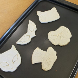 May include: Five unbaked sugar cookies on a black baking sheet. The cookies are shaped like a cat, a skull, a mask, a hat, and a cat mask.
