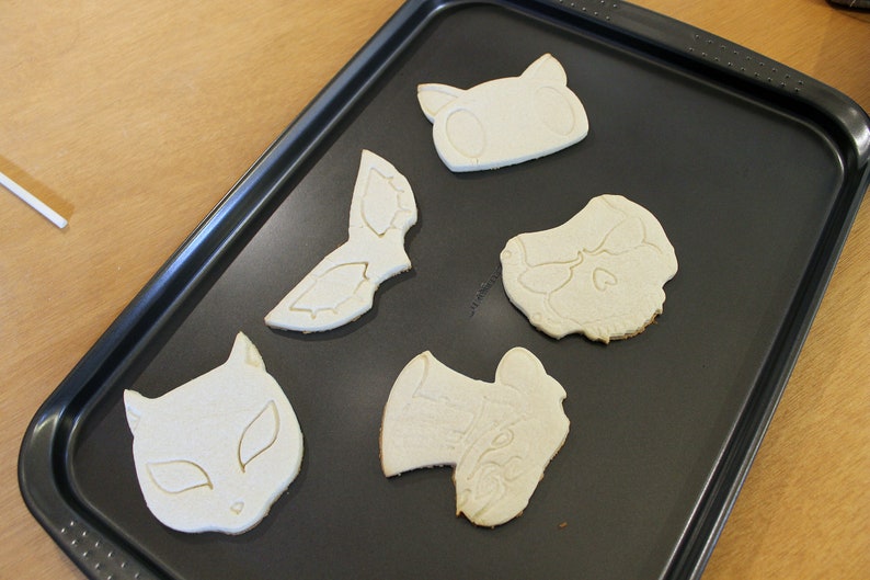 May include: Five unbaked sugar cookies on a black baking sheet. The cookies are shaped like a cat, a skull, a mask, a hat, and a cat mask.