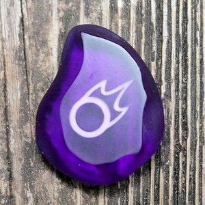 May include: A purple glass flame-shaped charm with a white symbol inside. The symbol is a circle with a flame coming out of the top.