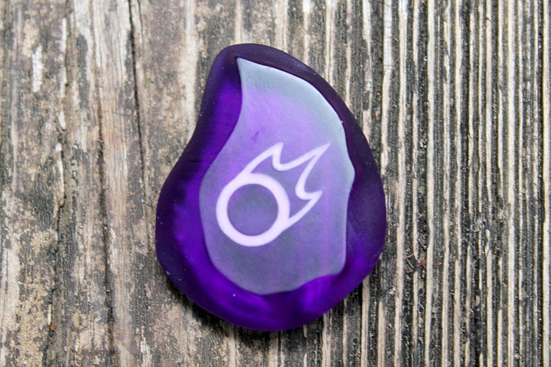 May include: A purple glass flame-shaped charm with a white symbol inside. The symbol is a circle with a flame coming out of the top.