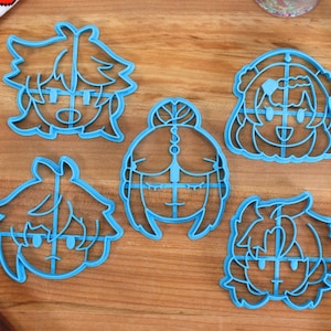 Puede incluir: Cinco cortadores de galletas de pl&aacute;stico azul con forma de caras de dibujos animados. Los cortadores est&aacute;n sobre una superficie de madera.