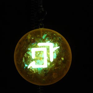 May include: A yellow glass orb with a white symbol inside. The orb is hanging from a black cord.
