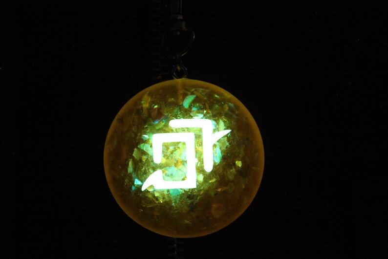 May include: A yellow glass orb with a white symbol inside. The orb is hanging from a black cord.