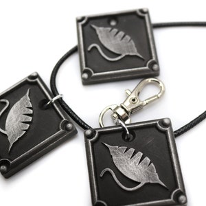 Puede incluir: Tres colgantes de metal plateados con fondo negro y un dise&ntilde;o de hoja plateada. Los colgantes est&aacute;n unidos a un cord&oacute;n negro con un cierre plateado.