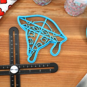 May include: A blue plastic cookie cutter shaped like a slice of pizza with pepperoni and cheese. The cutter is on a wooden surface with a ruler and a blue spatula.