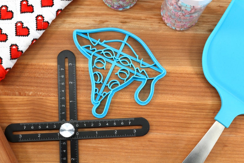 May include: A blue plastic cookie cutter shaped like a slice of pizza with pepperoni and cheese. The cutter is on a wooden surface with a ruler and a blue spatula.