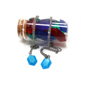 May include: Two necklaces with blue gem pendants hang from a glass bottle filled with colorful gem-shaped charms. The bottle has a cork stopper.