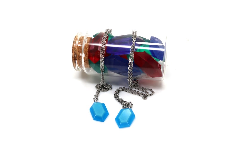 May include: Two necklaces with blue gem pendants hang from a glass bottle filled with colorful gem-shaped charms. The bottle has a cork stopper.