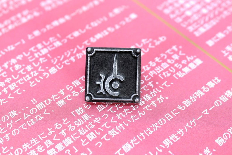 May include: A silver metal pin with a black background and a stylized symbol of a circle with a spike and a smaller circle inside. The symbol is surrounded by a border with spikes.