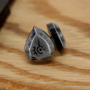 May include: A pair of silver teardrop-shaped earrings with a black symbol on each. The earrings are on a wooden surface.