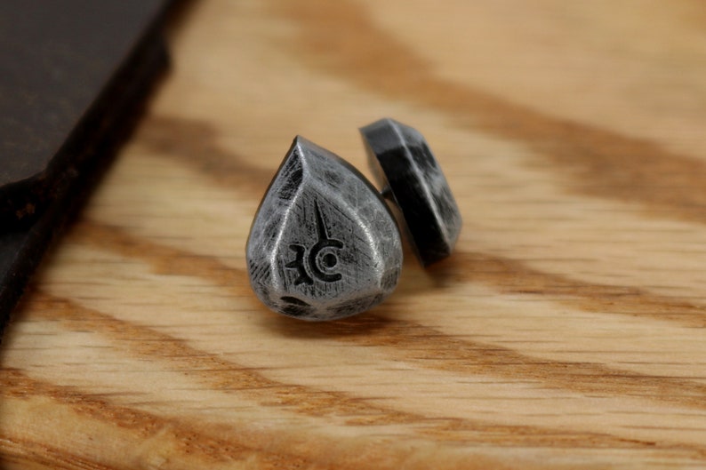 May include: A pair of silver teardrop-shaped earrings with a black symbol on each. The earrings are on a wooden surface.