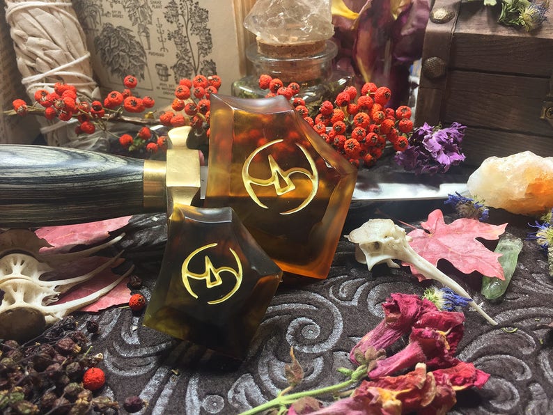 May include: Two amber-colored resin dice with gold-colored symbols on each face. The dice are on a dark brown surface with dried flowers and other natural elements.
