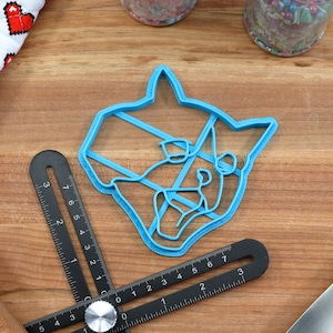 May include: A blue plastic cookie cutter shaped like a cat's head. The cutter is on a wooden surface with a ruler and a rolling pin.