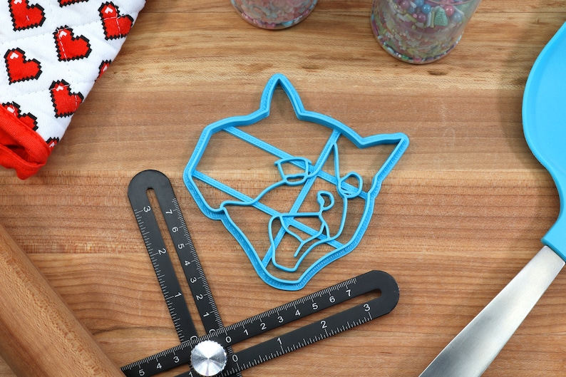 May include: A blue plastic cookie cutter shaped like a cat's head. The cutter is on a wooden surface with a ruler and a rolling pin.