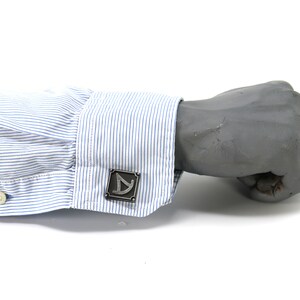 May include: A pair of silver cufflinks with a geometric design. The cufflinks are on a blue and white striped shirt cuff.