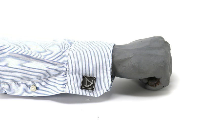 May include: A pair of silver cufflinks with a geometric design. The cufflinks are on a blue and white striped shirt cuff.