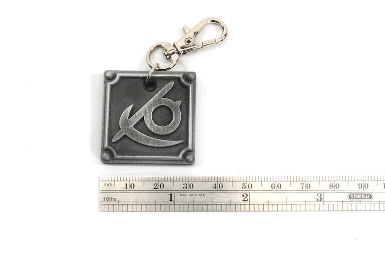 May include: A silver metal keychain with a square charm featuring a stylized eye symbol. The charm is about 3 cm wide.
