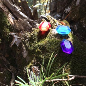 May include: Three plastic gems in red, blue, and purple, sitting on a mossy tree trunk in a forest setting.