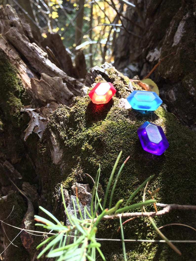 May include: Three plastic gems in red, blue, and purple, sitting on a mossy tree trunk in a forest setting.