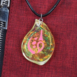 May include: A teardrop-shaped pendant necklace with a pink symbol inside. The pendant is made of clear iridescent glass and hangs from a black cord.