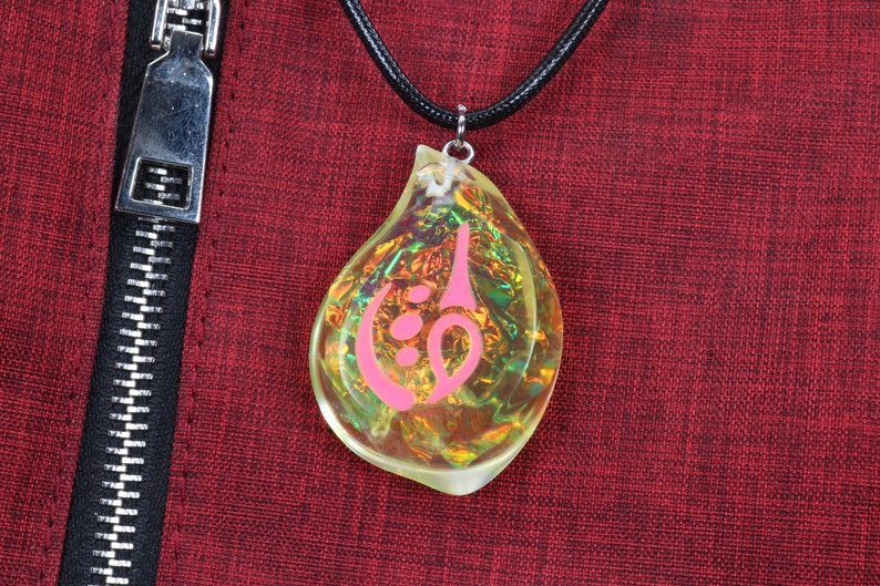 May include: A teardrop-shaped pendant necklace with a pink symbol inside. The pendant is made of clear iridescent glass and hangs from a black cord.