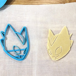 May include: A blue plastic cookie cutter and a light brown cookie in the shape of a cartoon character's head. The cookie cutter is on the left and the cookie is on the right.