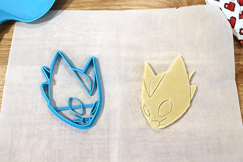 May include: A blue plastic cookie cutter and a light brown cookie in the shape of a cartoon character's head. The cookie cutter is on the left and the cookie is on the right.