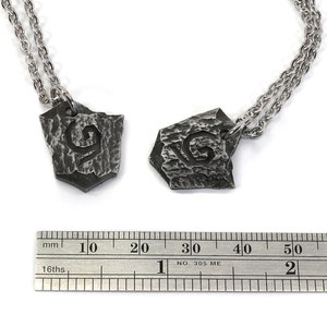 May include: Two silver necklaces with matching pendants. Each pendant is a shield-shaped piece of metal with a textured surface and the letter "G" engraved on it. The necklaces are shown next to a ruler, with the pendants measuring approximately 2 cm in length.