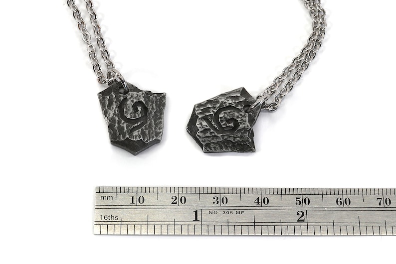 May include: Two silver necklaces with matching pendants. Each pendant is a shield-shaped piece of metal with a textured surface and the letter "G" engraved on it. The necklaces are shown next to a ruler, with the pendants measuring approximately 2 cm in length.