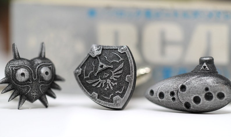May include: Three silver cufflinks featuring iconic symbols from the Legend of Zelda video game series: the Triforce, the Master Sword, and the Ocarina of Time.