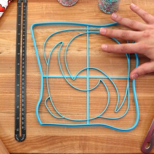 May include: A blue plastic mold for making toast with a swirl design. The mold is on a wooden cutting board with a ruler, rolling pin, and spatula.