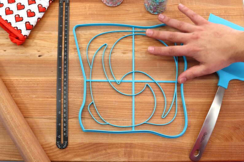 May include: A blue plastic mold for making toast with a swirl design. The mold is on a wooden cutting board with a ruler, rolling pin, and spatula.