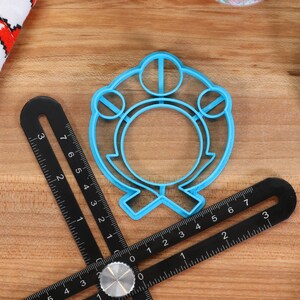 May include: A blue plastic cookie cutter shaped like a ring with three circles on top. The cutter is on a wooden surface with a ruler.