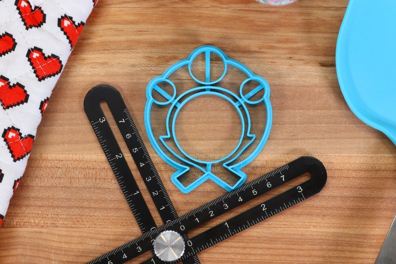 May include: A blue plastic cookie cutter shaped like a ring with three circles on top. The cutter is on a wooden surface with a ruler.
