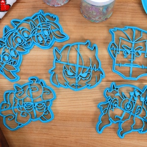 May include: Five blue plastic cookie cutters shaped like cartoon faces. The cutters are on a wooden surface.