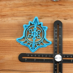 May include: A blue plastic snowflake cookie cutter with a detailed design. The cutter is on a wooden surface with a ruler next to it.
