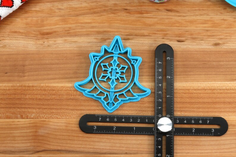 May include: A blue plastic snowflake cookie cutter with a detailed design. The cutter is on a wooden surface with a ruler next to it.